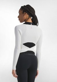 Langärmeliges Oberteil aus weißem und schwarzem Rippstoff, mit Ausschnitt am Rücken und tailliertem Schnitt. Kombiniert mit schwarzen Leggings.