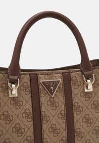 Sac à main Guess marron à motifs avec poignées en cuir, ferrures dorées, et une plaque triangulaire du logo Guess USA au centre devant.