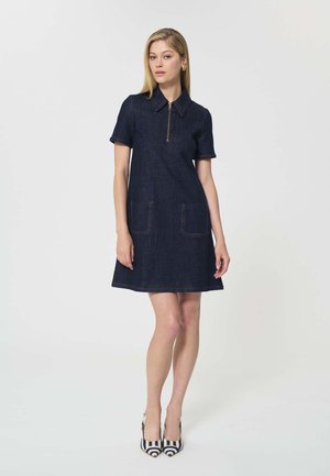Femme portant une robe courte en jean foncé à manches courtes avec des poches avant et un col zippé, associée à des escarpins à rayures noires et blanches.
