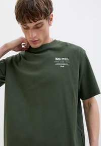 Olivegrøn bomulds t-shirt med korte ærmer, rund hals og hvid tekstgrafik på venstre bryst. Blød tekstur, afslappet pasform.