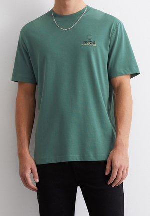 Camiseta de algodón verde con mangas cortas y cuello redondo. Presenta un pequeño diseño de un globo impreso y texto en la parte izquierda del pecho.