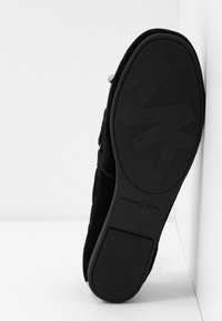 Semelle noire d'une chaussure Michael Kors avec logo MK en relief et talon texturé, inclinée contre une surface d'angle blanche.