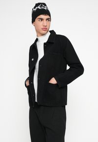 Čierna rifľová bunda s voľným strihom, s lesklým povrchom, zapínaním na gombíky, dvoma prednými vreckami a prešívanými detailmi. Kombinovaná s bielym rolákom.