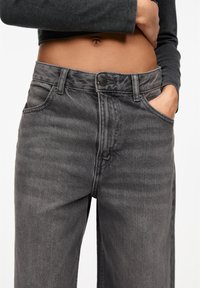 Milieu d'une personne portant un jean noir délavé taille haute, une main dans la poche et un haut court à manches longues foncé.