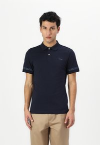 Tricou polo albastru închis cu guler, mâneci scurte și trei butoane. Logo-ul "GUESS" imprimat pe piept și pe marginea mânecii.