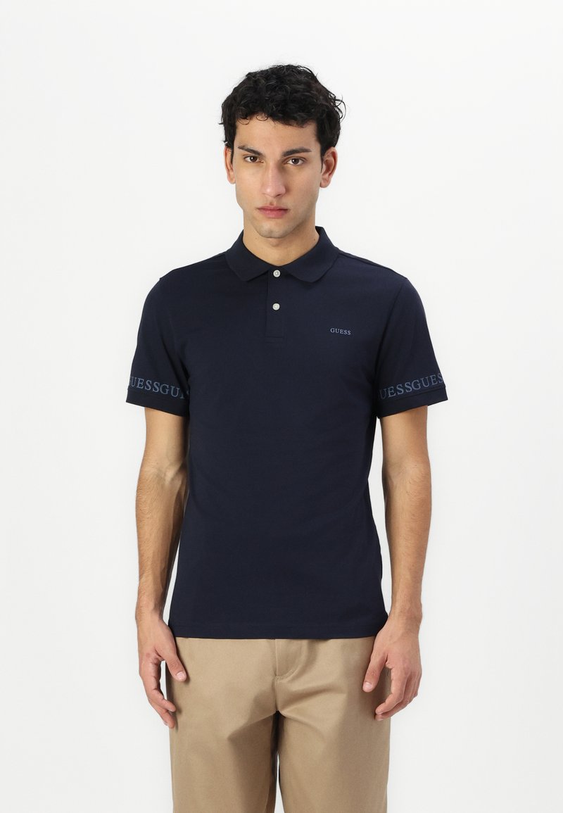 Tricou polo albastru închis cu guler, mâneci scurte și trei butoane. Logo-ul "GUESS" imprimat pe piept și pe marginea mânecii.