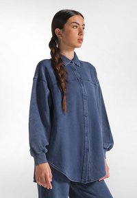 Camicia oversize blu con bottoni, realizzata in un tessuto morbido, dotata di colletto e maniche a palloncino, con orlo leggermente curvo e cuciture visibili.