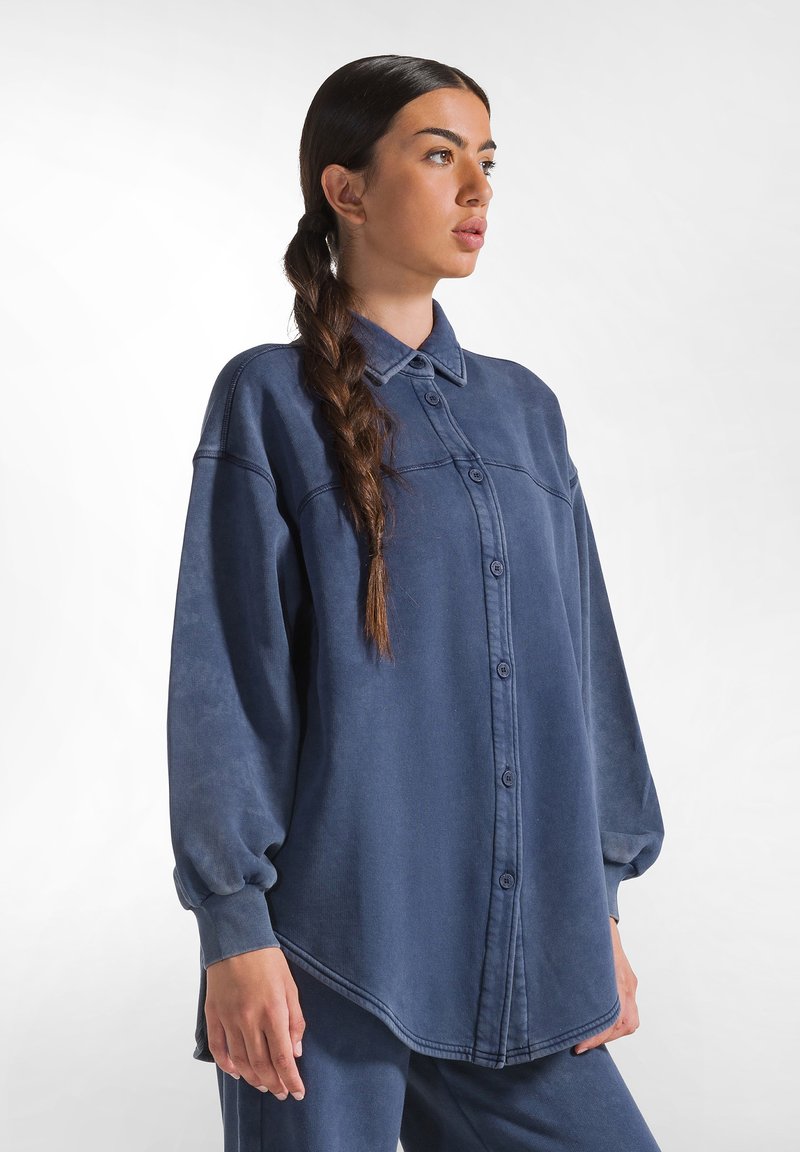 Camicia oversize blu con bottoni, realizzata in un tessuto morbido, dotata di colletto e maniche a palloncino, con orlo leggermente curvo e cuciture visibili.