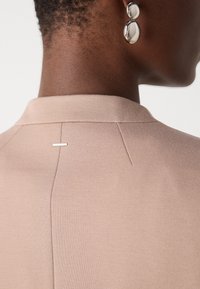 Gros plan sur le cou et l'épaule d'une personne portant un vêtement beige et une boucle d'oreille en argent composée de deux parties, suspendue à l'oreille.