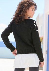 Schwarzer gestrickter Pullover mit geripptem Saum und Ärmeln, der weiße Akzentdetails an den Schultern aufweist. Über einer weißen Schicht unten getragen.