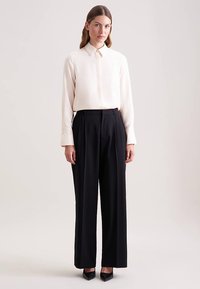 Camicia a maniche lunghe colore beige chiaro con colletto, abbinata a pantaloni neri a vita alta plissettati e décolleté nere con punta a punta. Texture liscia.