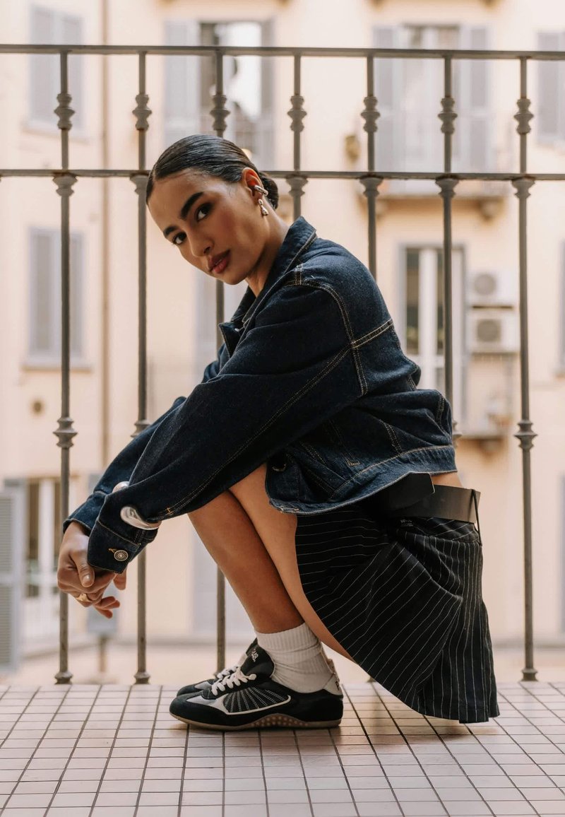 Jeune femme aux cheveux foncés portant une veste en jean, une jupe à fines rayures, des chaussettes blanches et des baskets, accroupie sur un balcon carrelé avec des balustrades en métal.
