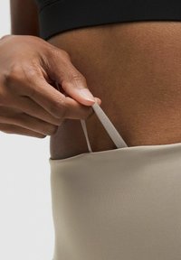 Beige stoffen hoge taille kledingstuk met een smalle elastische band. Een hand grijpt de band, waardoor de gladde textuur en het getailleerde ontwerp zichtbaar zijn.