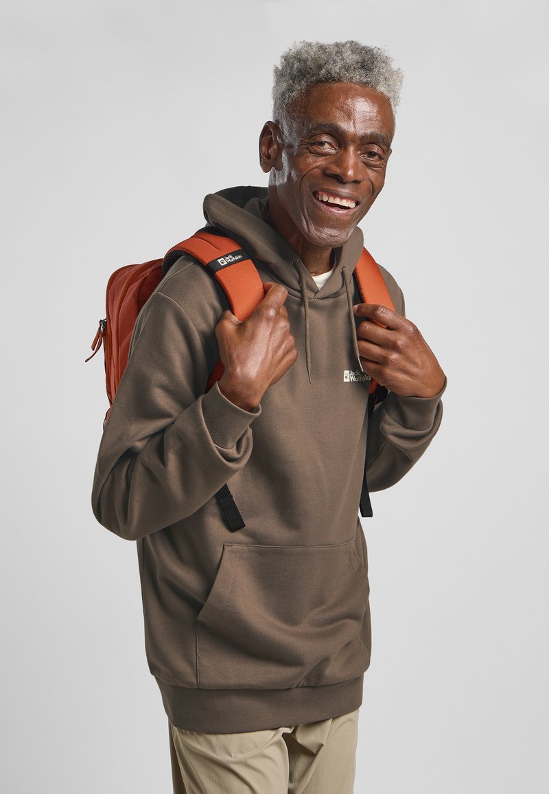 Bruine hoodie met een voorzak, gecombineerd met een oranje rugzak met gepolsterde schouderbanden en zwarte accenten. Gladde textuur.