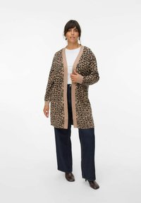 Cardigan léopard en brun et noir, avec une bordure beige. Présente des manches longues et un devant ouvert, associé à un jean large et foncé.