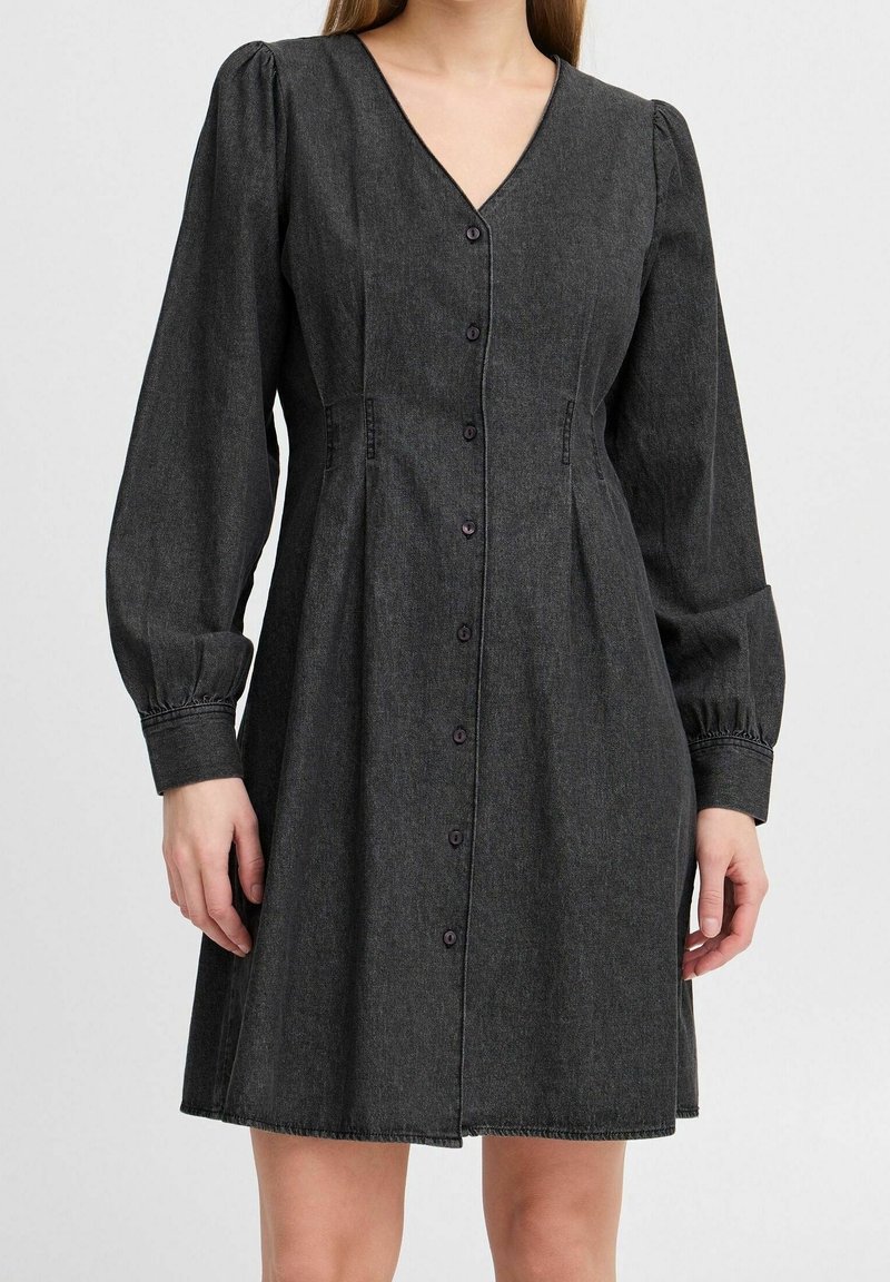 Robe en denim noir boutonnée, avec manches longues bouffantes et col en V, portée par une personne debout devant un fond uni.