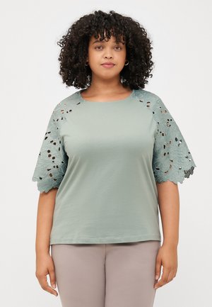 Model wearing a light green top with lace detail on sleeves, smiling, paired with light-colored pants against a plain background.