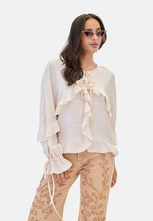 Mujer con blusa cremosa con volantes y pantalones beige bordados con hojas, combinada con gafas de sol de ojo de gato.