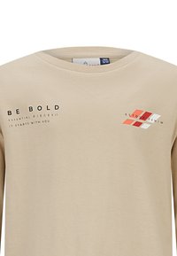 Lichtbeige langemouwen T-shirt met ronde hals, voorzien van zwarte tekst "BE BOLD" en "RETURN DENIM," plus rode en witte geometrische ontwerpelementen.