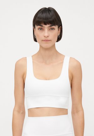 Woman with short dark hair wearing silver hoop earrings and a white crop top against a plain white background.