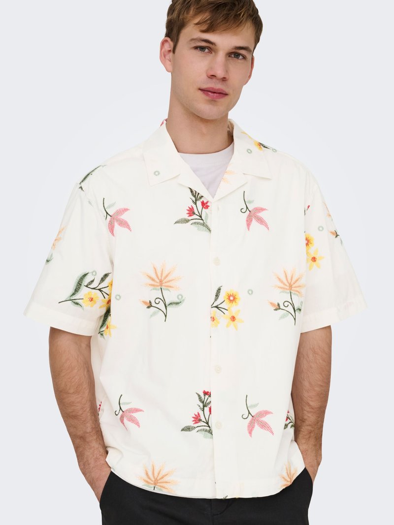 Jeune homme portant une chemise blanche à manches courtes avec des motifs floraux pêche, jaune et rouge, les mains dans les poches, debout devant un fond uni.