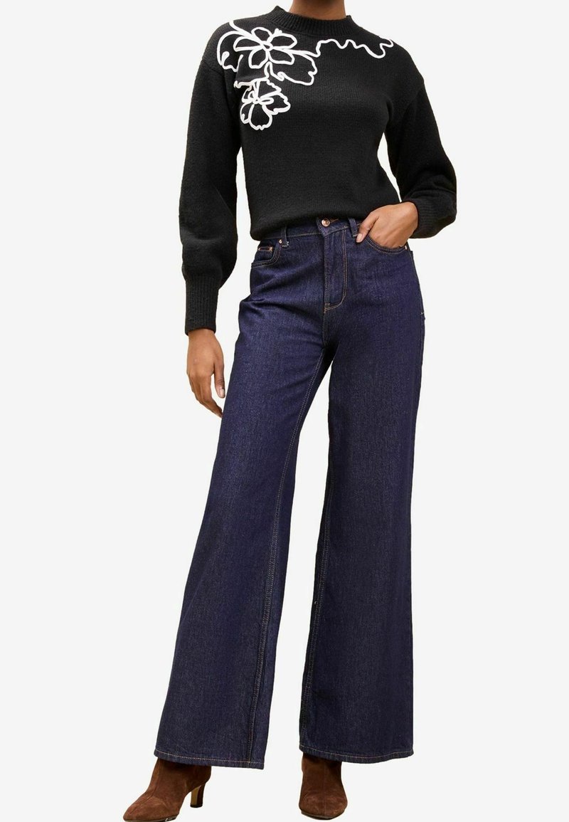Personne portant un pull noir avec une broderie florale blanche sur l'épaule, un jean large foncé et des bottes marron à talons, main dans la poche.
