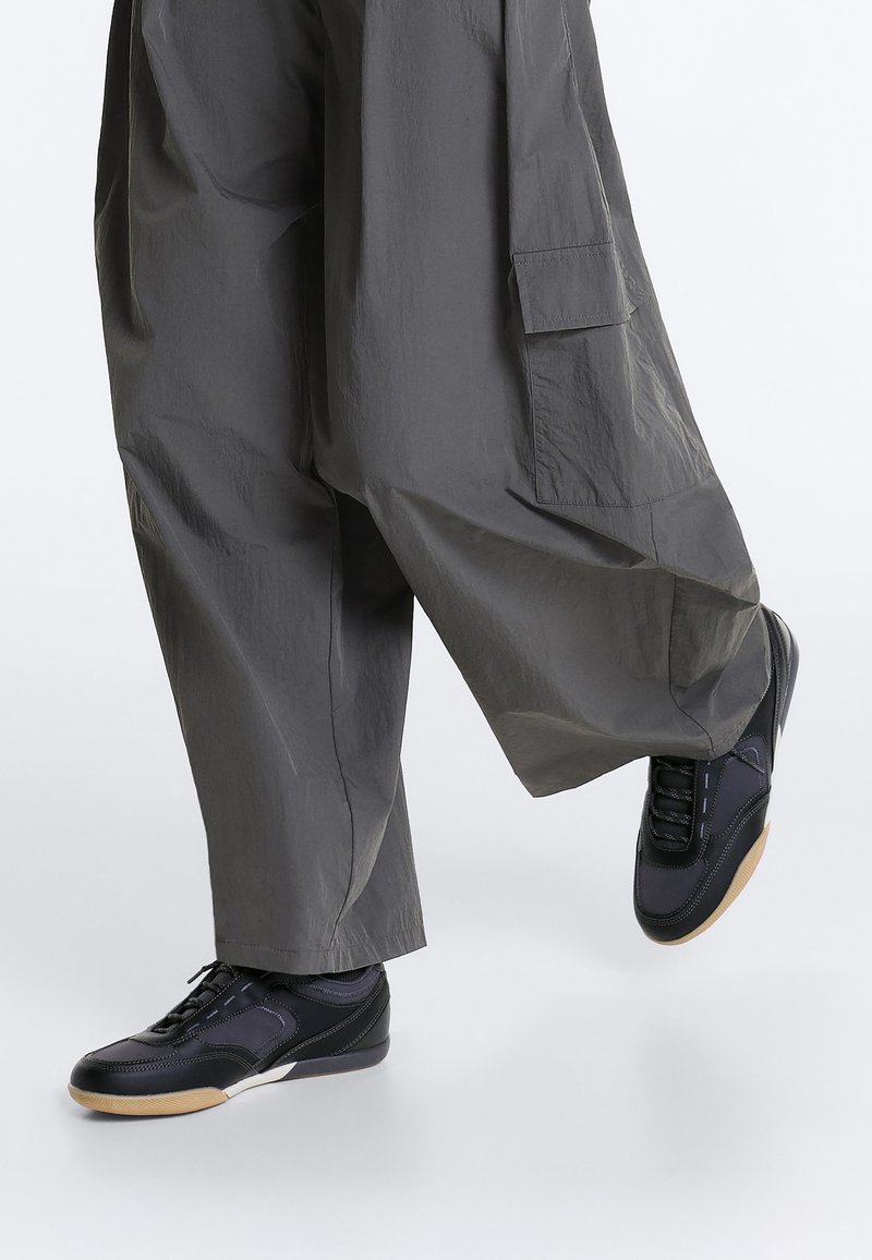 Pantalon gris foncé à jambes larges avec grande poche latérale et baskets basses noires à semelles en gomme sur une personne levant un pied.