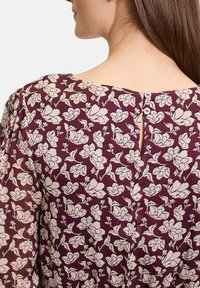 Bluse in Weinrot mit floralem Muster und einem Schlitz am Rücken, der mit zwei weißen Knöpfen verziert ist. Der Stoff hat eine strukturierte, leicht zerknitterte Optik.