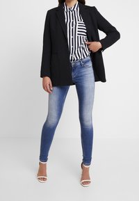 Blazer noir porté sur une chemise à rayures à boutons avec une poche, associé à un jean skinny bleu clair et des sandales à talons blanches.