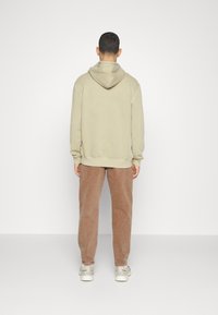 Beige huppari, jossa on nyöri, yhdistettynä ruskeisiin corduroy-housuihin. Malli seisoo selkä kameraan päin, kädet sivuilla, minimalistinen tausta.