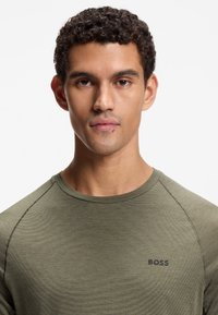 Oliwkowa, długorękawowa koszula o prążkowanej fakturze, z okrągłym dekoltem i czarnym logo "BOSS" na lewej piersi. Szwy zdobią rękawy.