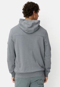 Grijze hoodie met een capuchon en trekkoord, geribbelde boorden en zoom, zacht materiaal en platte naden. Geen zichtbare logo's of patronen. Draagt bijpassende lichtgroene broek.