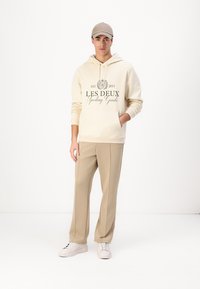 Beige hoodie with front pocket and logo, paired with beige tailored pants. Model wearing white sneakers and a beige cap.