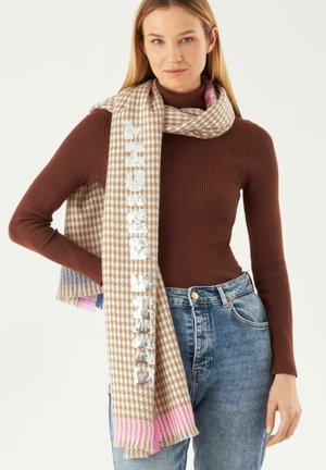 Écharpe marron à motif pied-de-poule avec des bords frangés roses et un texte en sequins argentés. Portée sur un haut marron ajusté et un jean taille haute bleu.