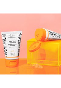 Jelly cleanser in a squeeze tube with a white base and black geometric patterns. Orange lid; product flows onto a yellow geometric block.