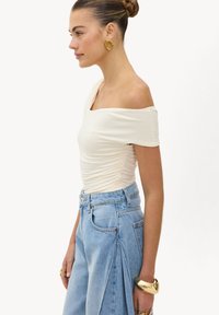 Femme avec les cheveux en chignon portant un haut blanc à épaules dénudées, un jean bleu plissé taille haute, de grandes boucles d'oreilles en or, un bracelet épais en or et des bagues.