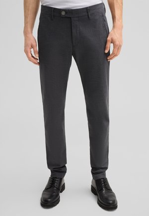 Pantalon taillé gris foncé avec un motif texturé subtil, design à jambes droites, fermer par un bouton à l'avant, et poches latérales, associé à des chaussures noires.