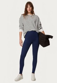 Maglietta a maniche lunghe a righe bianche e nere, leggings blu e scarpe da ginnastica bianche. La modella tiene in mano una giacca trapuntata nera. Design casual e aderente.