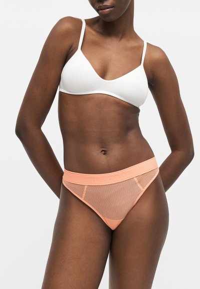 Γυναίκα με λευκό μπραλέτο και πετρόλ ριμπειδωτό εσώρουχο μπικίνι σε απλό ανοιχτό φόντο.