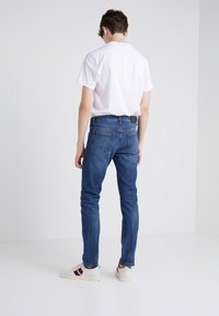 Blå smala jeans i denim, med bakfickor och en svart läderlapp vid midjan. Kombineras med en vit t-shirt och sneakers.