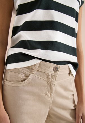 Personne portant un pantalon beige et une chemise rayée horizontale noire et blanche, les mains détendues sur les côtés.