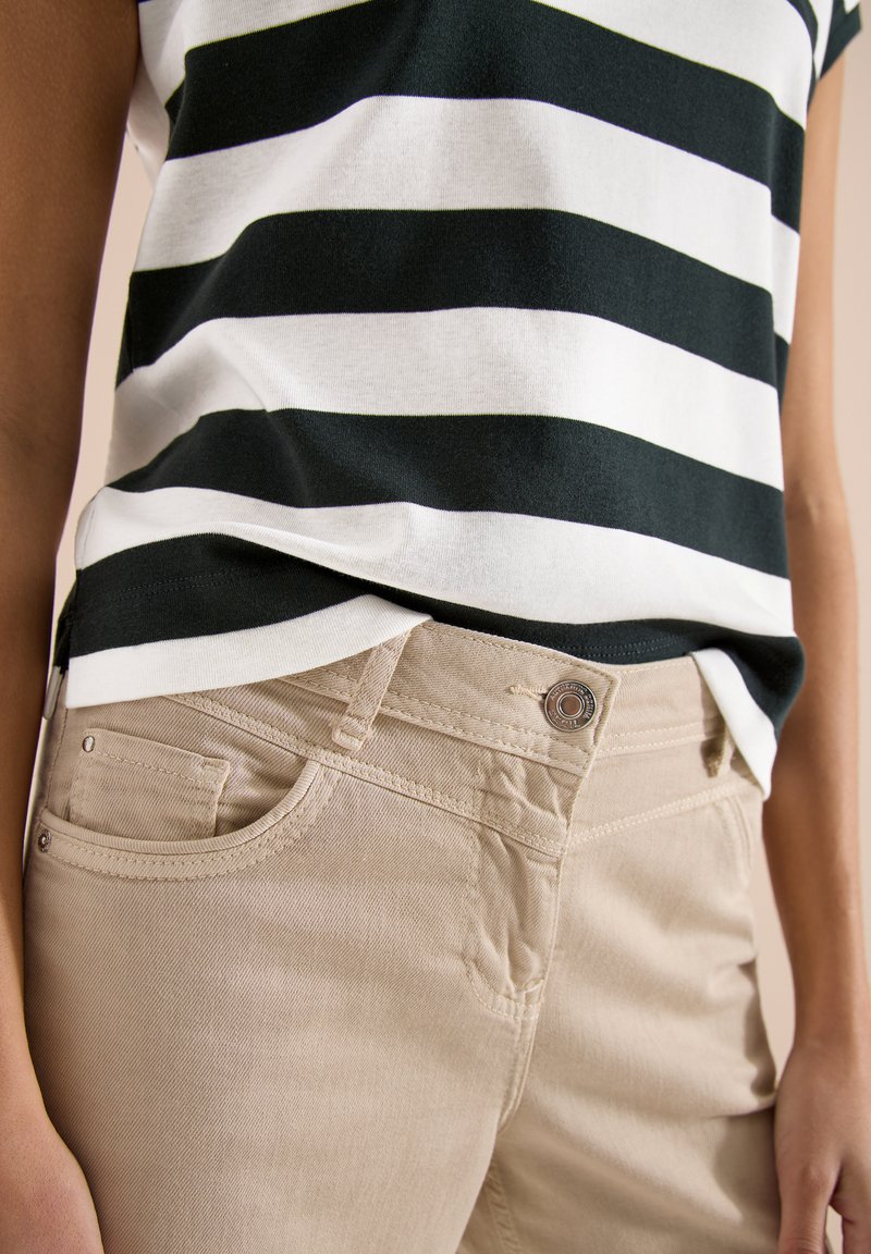 Personne portant un pantalon beige et une chemise rayée horizontale noire et blanche, les mains détendues sur les côtés.