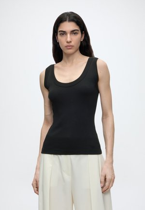 Femme aux longs cheveux noirs portant un haut noir sans manches et un pantalon blanc à jambes larges, debout devant un fond clair uni.