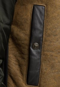 Bruin suede stof met een textuurafwerking, voorzien van een zwarte leren accent en een cirkelvormige metalen logo badge. Gestikte randen zichtbaar.