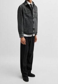 Giacca in denim nera con due tasche sul petto, chiusura a bottoni e vestibilità rilassata, indossata sopra una camicia bianca e abbinata a pantaloni e scarpe neri.