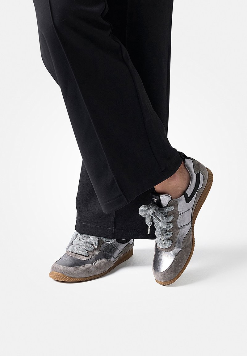 Personne portant un pantalon noir et des baskets métalliques gris avec des semelles en gomme, pieds croisés sur fond blanc.