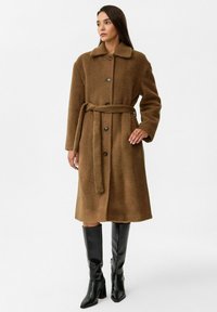 Bruine wollen jas met een kraag, knopen aan de voorkant en een strikriem. Heeft lange mouwen en een middellange snit, gestyled met kniehoge zwarte laarzen.