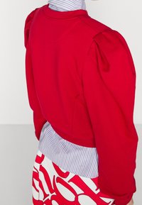 Sudadera roja con hombros abullonados, que presenta un cuello a rayas y una cinturilla ancha. La parte inferior tiene un patrón abstracto audaz en blanco y rojo.