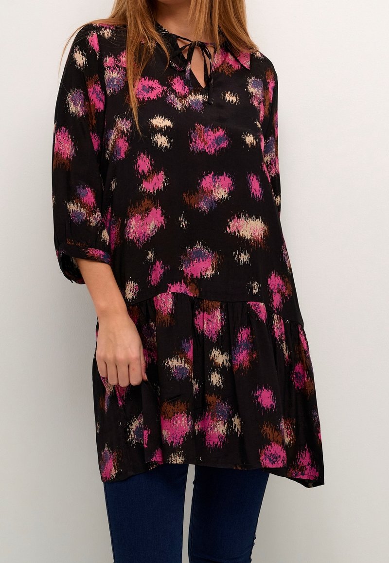 Femme portant une tunique noire avec des taches abstraites roses, marron et beige et un lien à l'encolure, associée à un pantalon bleu foncé.
