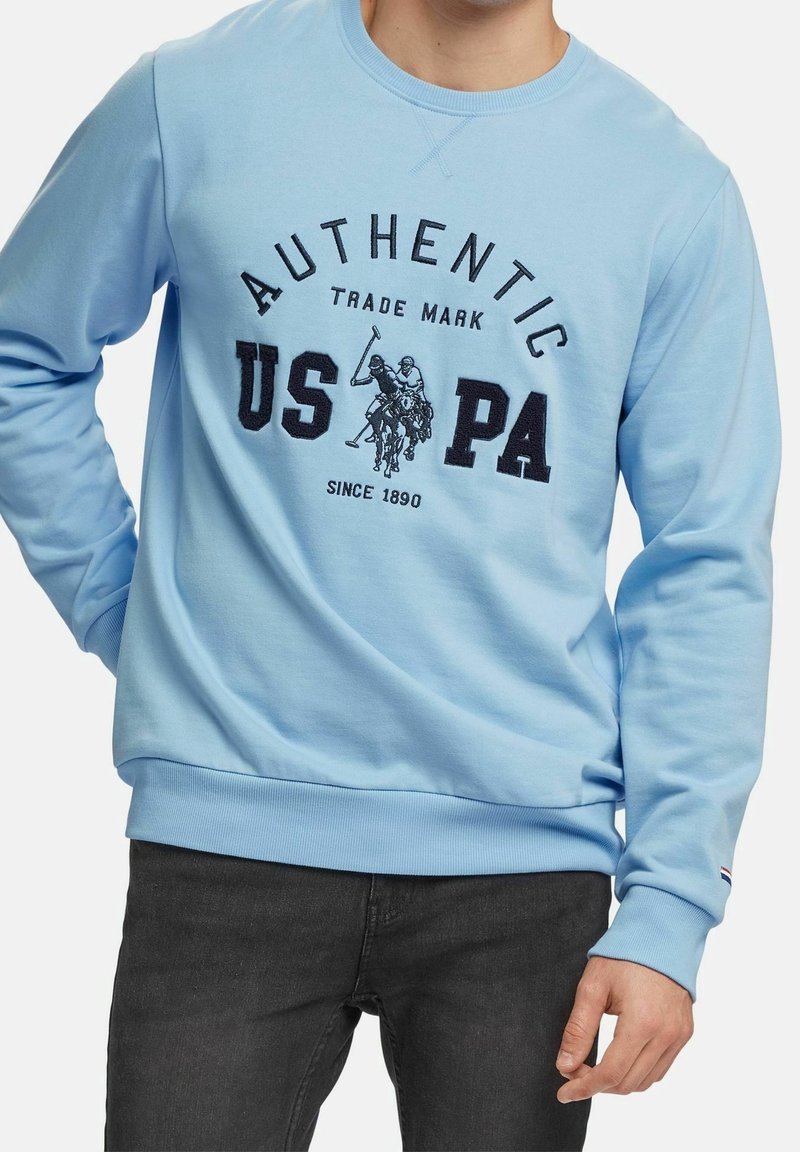 Sweat-shirt bleu clair avec texte noir « AUTHENTIC USPA » et deux joueurs de polo brodés à l'avant, porté par une personne en jean noir.
